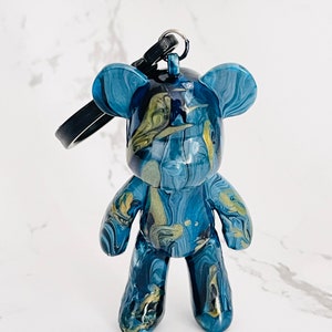 Pode incluir: Um chaveiro de urso de pel&uacute;cia azul e dourado marmorizado. O pingente em forma de urso tem um acabamento brilhante, uma al&ccedil;a preta e um chaveiro. O padr&atilde;o de cores se assemelha &agrave; tinta fluida.