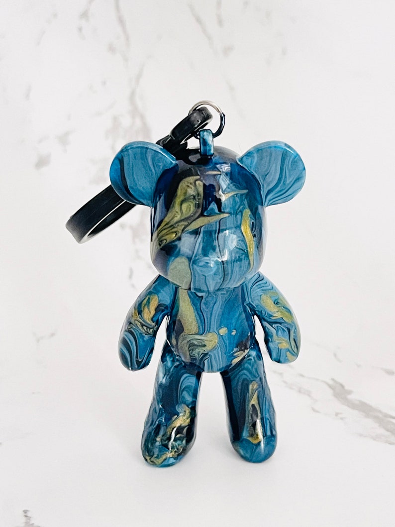 Pode incluir: Um chaveiro de urso de pel&uacute;cia azul e dourado marmorizado. O pingente em forma de urso tem um acabamento brilhante, uma al&ccedil;a preta e um chaveiro. O padr&atilde;o de cores se assemelha &agrave; tinta fluida.