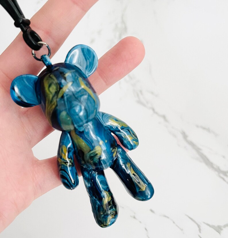 Pode incluir: Pingente de urso de pel&uacute;cia azul e dourado marmorizado com um cord&atilde;o preto. O urso tem um acabamento brilhante e um padr&atilde;o &uacute;nico. O pingente &eacute; um acess&oacute;rio decorativo.