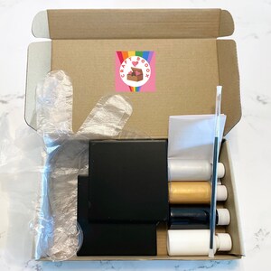 Peut inclure: Une bo&icirc;te en carton ouverte remplie de fournitures artisanales. La bo&icirc;te contient une toile noire, quatre bouteilles de peinture, un pinceau et une paire de gants en plastique. La bo&icirc;te est &eacute;tiquet&eacute;e "Craft Boox".