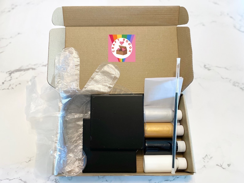 Peut inclure: Une bo&icirc;te en carton ouverte remplie de fournitures artisanales. La bo&icirc;te contient une toile noire, quatre bouteilles de peinture, un pinceau et une paire de gants en plastique. La bo&icirc;te est &eacute;tiquet&eacute;e "Craft Boox".
