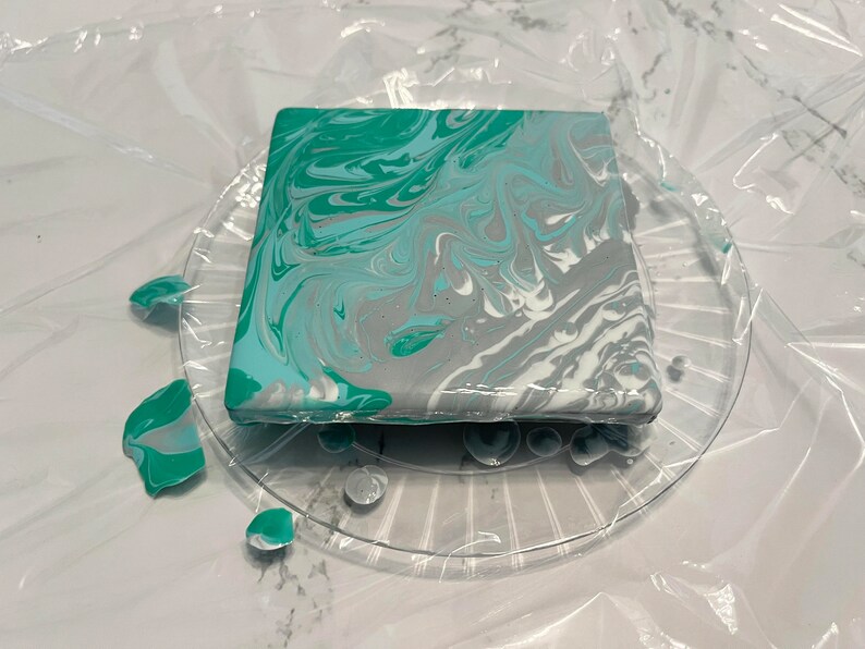 Peut inclure: Un dessous de verre carr&eacute; avec un motif marbr&eacute; dans des tons de turquoise, gris et blanc. Le dessous de verre est pos&eacute; sur une assiette en plastique transparente.