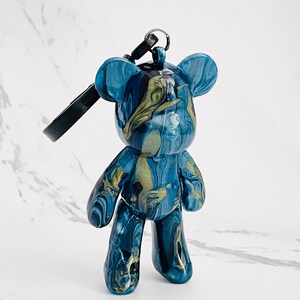 Pode incluir: Uma estatueta de urso de peluche azul e dourado marmoreado com um porta-chaves. O urso tem um acabamento brilhante e um padr&atilde;o &uacute;nico de redemoinho de azul escuro, azul claro e dourado. O porta-chaves &eacute; preto.