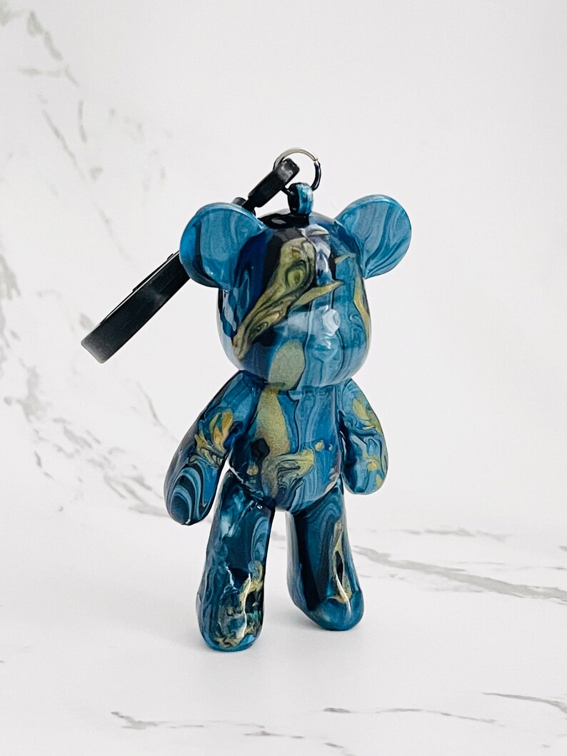 Pode incluir: Uma estatueta de urso de peluche azul e dourado marmoreado com um porta-chaves. O urso tem um acabamento brilhante e um padr&atilde;o &uacute;nico de redemoinho de azul escuro, azul claro e dourado. O porta-chaves &eacute; preto.