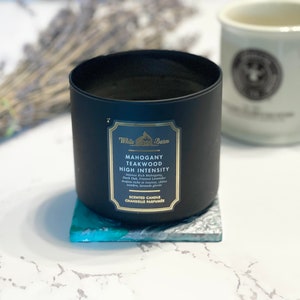 Peut inclure: Un bocal de bougie noir avec une &eacute;tiquette dor&eacute;e qui indique "Mahogany Teakwood High Intensity" avec des notes de "Mystic Pink Mahogany, Dark Oak, Frosted Lavender, Amber Musk, Cedarwood, Clove, Sandalwood, Brandy, Ginger". La bougie est pos&eacute;e sur un dessous-de-plat bleu et blanc.