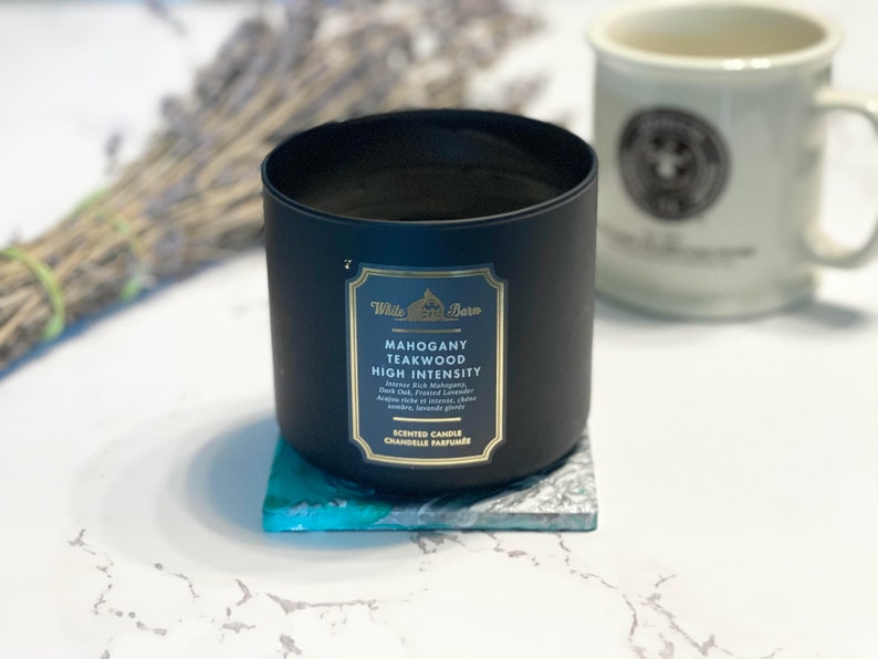 Peut inclure: Un bocal de bougie noir avec une &eacute;tiquette dor&eacute;e qui indique "Mahogany Teakwood High Intensity" avec des notes de "Mystic Pink Mahogany, Dark Oak, Frosted Lavender, Amber Musk, Cedarwood, Clove, Sandalwood, Brandy, Ginger". La bougie est pos&eacute;e sur un dessous-de-plat bleu et blanc.