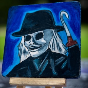 May include: A square painting on a small wooden easel featuring a stylized character with a white skull-like face, wearing a black fedora and a dark coat. The character has long white hair and holds a silver hook tool against a vibrant blue background.