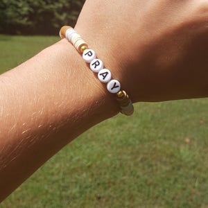 May include: A white beaded bracelet with gold beads and the word "PRAY" spelled out in black letters.