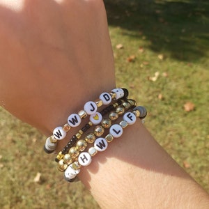 May include: A stack of beaded bracelets with white letter beads spelling out "WWJD" and "HWLF" with a gold heart bead between the words. The bracelets are on a person's wrist.