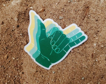 May include: A green, yellow, and blue hand gesture sticker with a shaka sign. The sticker is on a brown sandy surface.