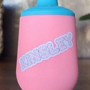 Pu&ograve; includere: Borraccia termica rosa e blu con un adesivo bianco che dice "Kinsley" in un font con motivo.