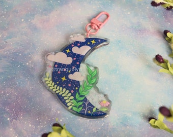 Può includere: Un ciondolo a forma di mezzaluna blu con una nuvola rosa e bianca, foglie verdi e stelle dorate. Il ciondolo è attaccato a un portachiavi in metallo rosa. Il ciondolo è su uno sfondo blu chiaro con macchie bianche.