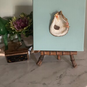 May include: A decorative piece featuring a light blue square canvas with an oyster shell affixed to it. The shell has a gold-colored trim and is mounted on a small bamboo easel. The background includes a marble surface and decorative items.