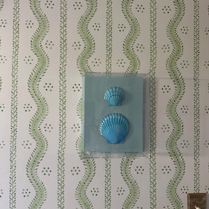 May include: Two bright blue seashell decorations are mounted on a light blue background, encased in a clear frame. The seashells are set against a wallpaper with a white background and a green, wavy vine pattern.