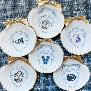 May include: Six white scallop shells with gold-painted edges, each featuring a different university logo in blue and white. The logos are displayed within a shield design, with decorative elements. The shells are arranged on a blue patterned surface.