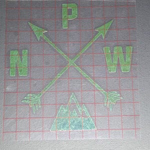 May include: A green vinyl decal featuring two crossed arrows, the letters P, N, and W, and a mountain graphic. The design is printed on a clear transfer sheet with a red grid pattern, intended for application onto a surface as a decorative element.