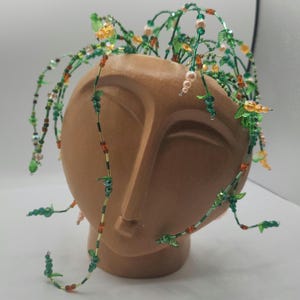 Peut inclure: Un cache-pot en céramique couleur terre cuite représentant un visage abstrait aux traits lisses. Le sommet est orné de multiples fils métalliques décorés de petites feuilles en verre vert, de perles orange translucides et de perles rose pâle.