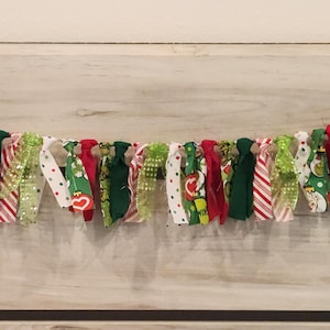 May include: A festive fabric rag garland featuring a variety of knotted strips in shades of red, lime green, and white. The fabric patterns include solid colors, candy cane stripes, polka dots, and illustrations of a green cartoon character.