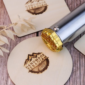 Puede incluir: Primer plano de un hierro de marcar metálico con una cabeza dorada. El hierro de marcar se utiliza para crear un logotipo personalizado en posavasos de madera. El logotipo presenta una cordillera y un diseño de anillos de árbol con el texto "HANDMADE ZEBADIAH".