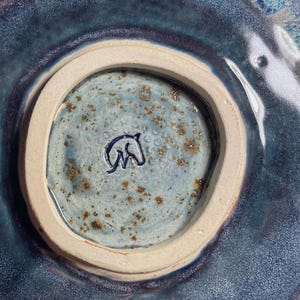 May include: The underside of a ceramic bowl featuring a circular foot ring. The centre is glazed in a speckled light blue with a dark blue stamped horse head logo. The surrounding rim is a deep, glossy blue with purple and black undertones.