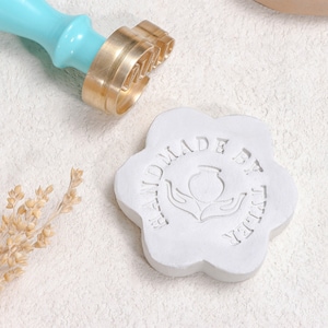 Puede incluir: Un sello de lacre turquesa y dorado con la palabra "LOVE" y un sello de cera blanco en forma de flor con el texto "HANDMADE BY TYLER". El sello tiene un mango turquesa.