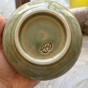 以下が含まれることがあります： セージグリーンのひび割れた釉薬仕上げが施された丸い陶器のボウル。底面には釉薬のかかっていないベージュ色の縁取りがある円形の高台があり、円形の枠の中にKHの文字が刻印されたメーカーのマークが施されています。