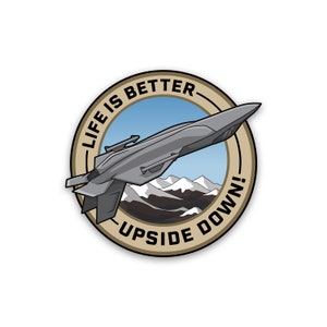 May include: A round sticker with a tan border and a blue sky background. The sticker features a cartoon jet flying upside down over a mountain range. The text "Life is better upside down!" is written around the edge of the sticker.