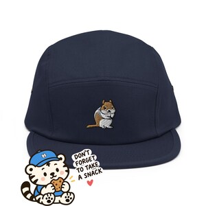 Peut inclure: Une casquette cinq pans bleu marine avec un écureuil brodé marron et blanc sur le devant. Dans le coin inférieur gauche, un tigre de dessin animé portant une casquette bleue mange un biscuit avec le texte "DON'T FORGET TO TAKE A SNACK" et un petit cœur rouge.