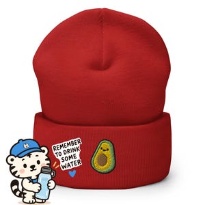 May include: A solid red cuffed beanie featuring an embroidered graphic of a smiling avocado, a small blue heart, and the text "REMEMBER TO DRINK SOME WATER" next to a cartoon tiger character holding a blue water bottle in a blue baseball cap.