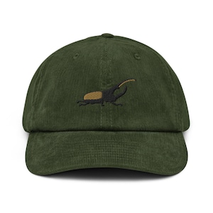 Peut inclure: Une casquette de baseball en velours côtelé vert olive foncé avec une broderie représentant un scarabée rhinocéros sur le devant. L'insecte est réalisé avec des fils noirs et beiges, mettant en valeur sa corne et son corps texturé.