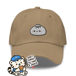 Op de afbeelding: Een beige honkbalpet met een geborduurde witte knoedelfiguur met een lachend gezicht en roze wangen. Een cartoon-tijger met een blauwe pet eet een koekje naast de tekst: DON'T FORGET TO TAKE A SNACK.