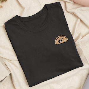 Könnte beinhalten: Ein schwarzes T-Shirt mit Rundhalsausschnitt und einer kleinen Stickerei auf der linken Brust. Das Motiv zeigt ein beigefarbenes, cartoonartiges Teigtäschchen mit lächelndem Gesicht, rosa Wangen und einer kleinen Hand, die ein winziges silbernes Teigtäschchen hält.