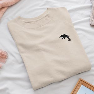 May include: A beige crewneck sweatshirt featuring a small, embroidered black and white orca whale graphic positioned on the upper left chest area. The garment has long sleeves, a ribbed neckline, and a simple, clean design with no other patterns.