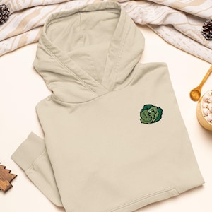Op de afbeelding: Een beige hoodie met een kleine, geborduurde groene koolafbeelding op de linkerborst. De trui met capuchon is voorzien van een kangoeroezak aan de voorzijde, lange mouwen met geribbelde boorden en een capuchon zonder trekkoord.