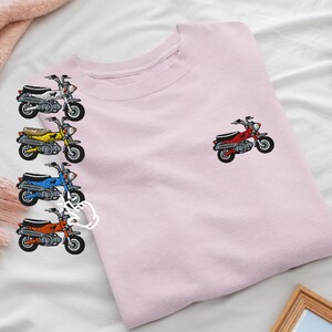 以下が含まれることがあります： 白、黄色、青、オレンジ色の刺繍入りミニチュアバイクが特徴の薄いピンク色のスウェットシャツ。右胸上部には赤いバイクが1台刺繍されています。バイクには黒いシートとタイヤが付いています。