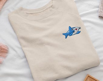 May include: A beige crewneck sweatshirt featuring a small, embroidered blue hammerhead shark on the upper left chest. The shark design includes white and dark blue details with a smiling expression, stitched onto the solid light-colored fabric.