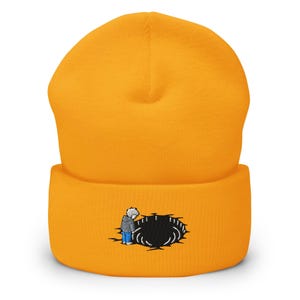 May include: A bright orange cuffed beanie featuring an embroidered graphic on the front. The design depicts a small figure in a grey hooded jacket and blue pants standing at the edge of a jagged, circular black hole with white outline details.