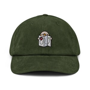 May include: A dark olive green corduroy baseball cap featuring an embroidered graphic of a small, light-coloured bird holding an open white map. The map displays a dotted path and a red location pin marker, centred on the front of the cap.