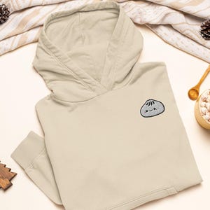 May include: A beige hooded sweatshirt featuring an embroidered graphic of a small, smiling grey dumpling on the left chest. The hoodie has long sleeves with ribbed cuffs, a front kangaroo pocket, and a hood with a visible seam detail at the neck.
