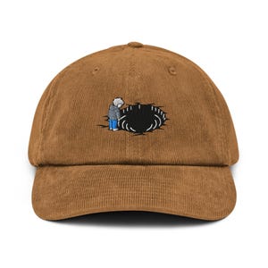 May include: A brown corduroy baseball cap with a curved brim. The cap features an embroidered design of a person looking into a black hole. The person is wearing a gray jacket and blue pants.
