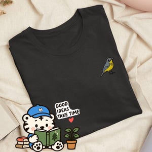 Puede incluir: Una camiseta negra con un pequeño pájaro amarillo y gris bordado en el pecho. Debajo del pájaro, un tigre de dibujos animados con gorra azul lee un libro verde junto a una planta en maceta y el texto GOOD IDEAS TAKE TIME con un corazón rojo.