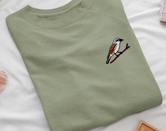 May include: A sage green crewneck sweatshirt featuring a small embroidered graphic of a bird with a grey head, brown wings, and a black eye stripe, perched on a brown branch on the upper right side of the chest area.