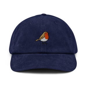 Op de afbeelding: Een marineblauwe baseballpet van corduroy met een geborduurde roodborst op het voorpaneel. De vogel is afgebeeld met een feloranje borst, bruine vleugels en een witte buik, staand op dunne zwarte pootjes tegen de donkerblauwe stof aan.