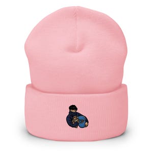 May include: A light pink beanie hat with a folded brim. The hat features an embroidered design of a person holding a teddy bear and a blue bowl. The beanie is made of a soft, knitted material.
