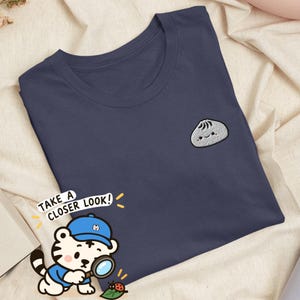 Peut inclure: Un t-shirt bleu marine foncé avec une petite broderie représentant une boulette blanche au visage souriant sur la poitrine gauche. Un tigre de dessin animé portant une casquette bleue et une loupe avec le texte TAKE A CLOSER LOOK! est présent.
