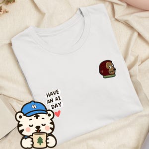 May include: A white t-shirt featuring a small embroidered brown monkey head on the upper right chest. Below the embroidery, the text "HAVE AN A1 DAY" is printed in black, accompanied by a small red heart and a cartoon tiger wearing a blue cap.
