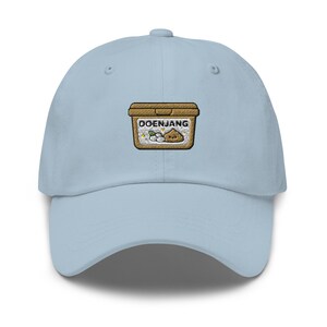 May include: A light blue baseball cap with an embroidered patch featuring the word "DOENJANG" above an illustration of a container with ingredients. The cap is made of a soft fabric and has a curved brim.
