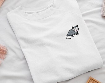 May include: A white long-sleeve sweatshirt featuring a small embroidered graphic of a gray and white opossum on the upper left chest area. The garment has a ribbed crew neckline and raglan sleeves, constructed from a solid white fabric.