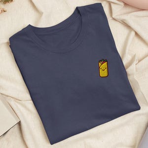 May include: A dark navy blue crew neck t-shirt featuring a small, embroidered graphic on the left chest. The embroidery depicts a yellow cartoon burrito with a smiling face, brown filling, and green toppings, rendered in a simple, clean style.