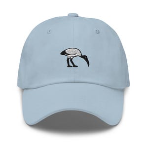 May include: Pale blue baseball cap featuring a black and white embroidered ibis design. The cap has a curved peak and an adjustable strap. The ibis is a stylized bird.